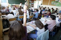 A classroom full of students listens to a teacher in front of a chalkboard. Two students' hands are raised.