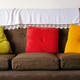 A brown couch with three throw pillows—one green, one red, one yellow—and a white blanket on the back