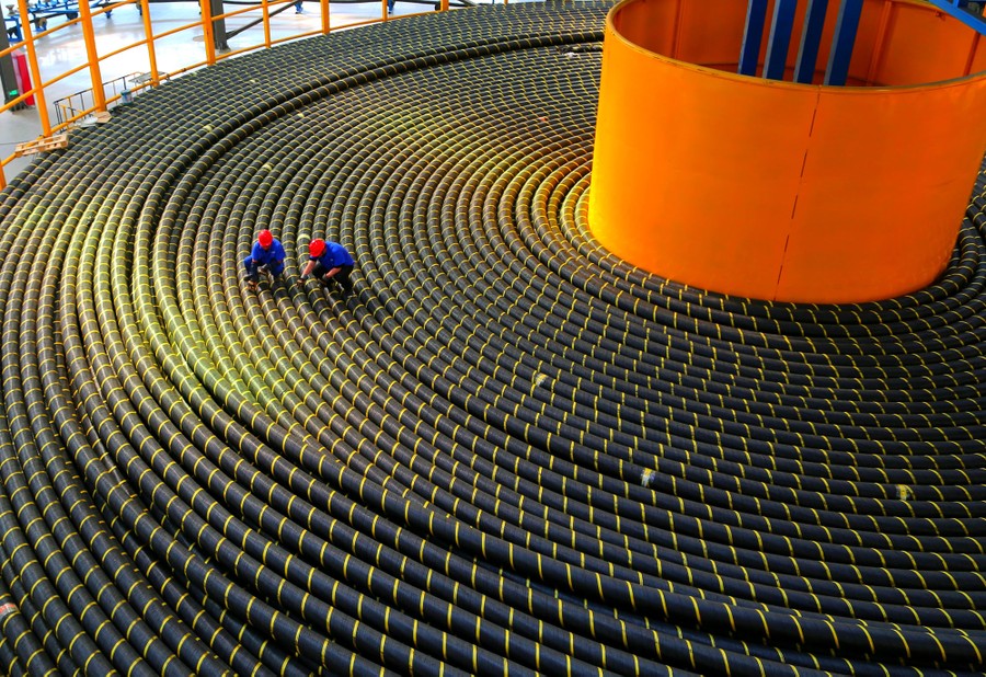 Two people kneel to examine part of a very long coil of thick cables.