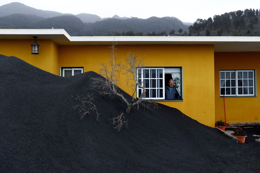 A man stands in a window of a house that is half-buried in dark-colored volcanic ash.