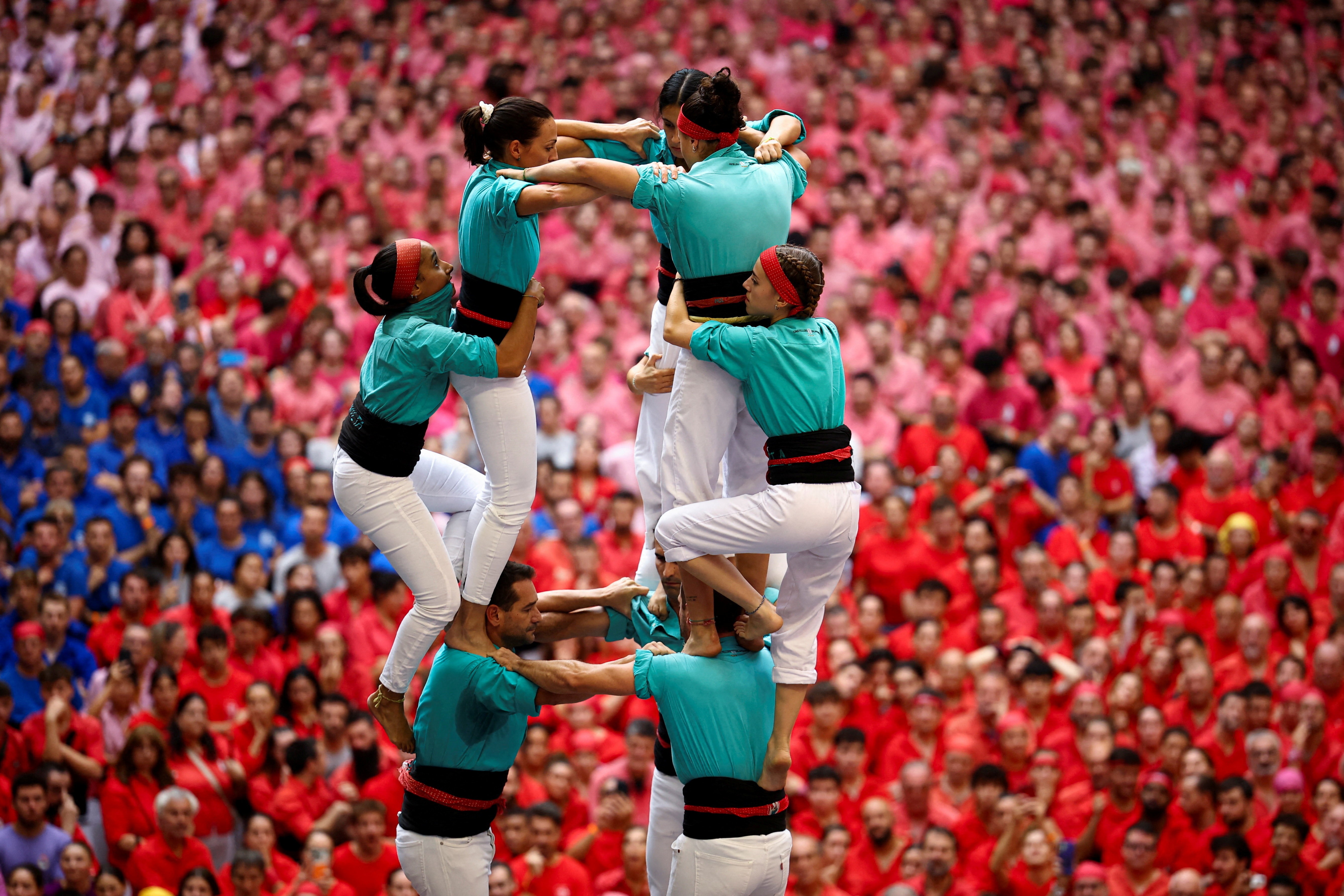 A zoomed-in view of several people climbing to the top of a human tower, with a large crowd of other teams visible in the background