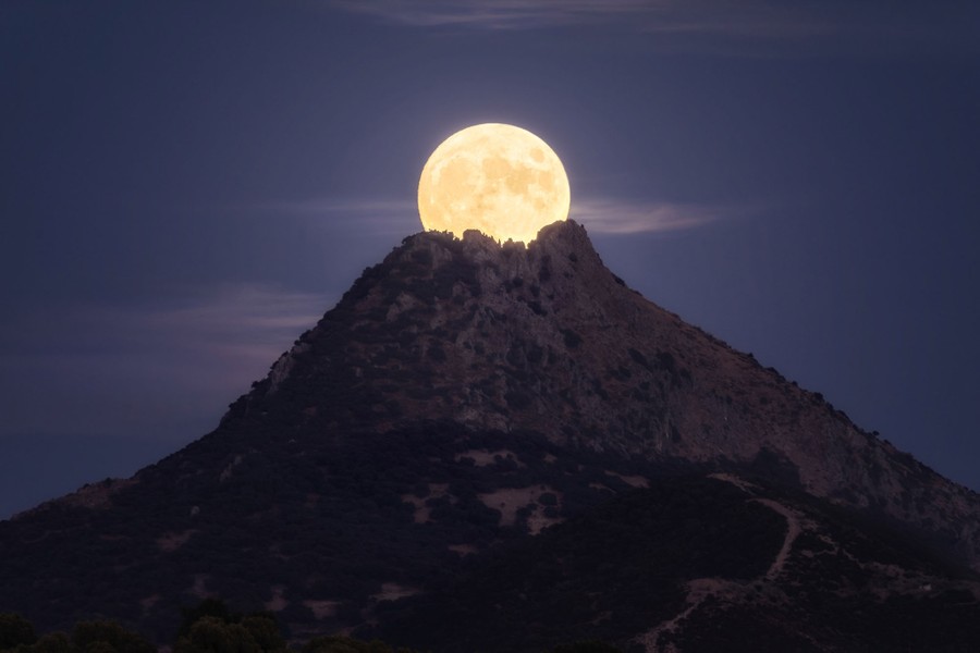 A photograph of the moon behind a mountain top, appearing as if the moon is emerging from the crater of a volcano.