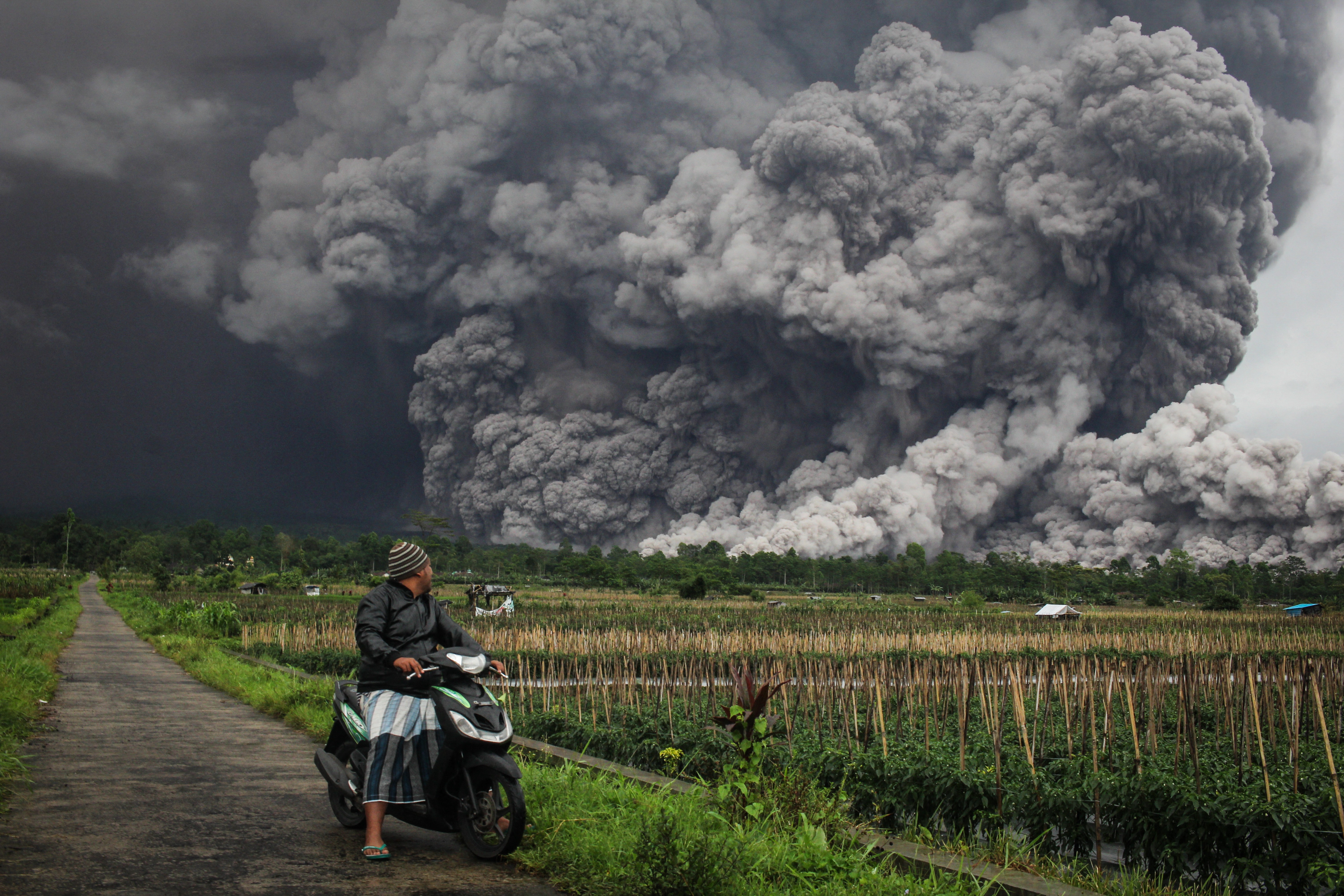 A man on a scooter looks at a huge cloud of volcanic ash billowing across a field behind him.