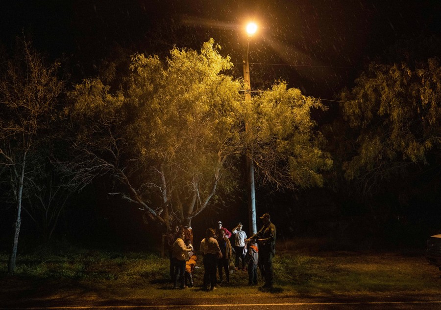 A U.S. Border Patrol agent speaks to a small group of migrants at night, beneath a tree.