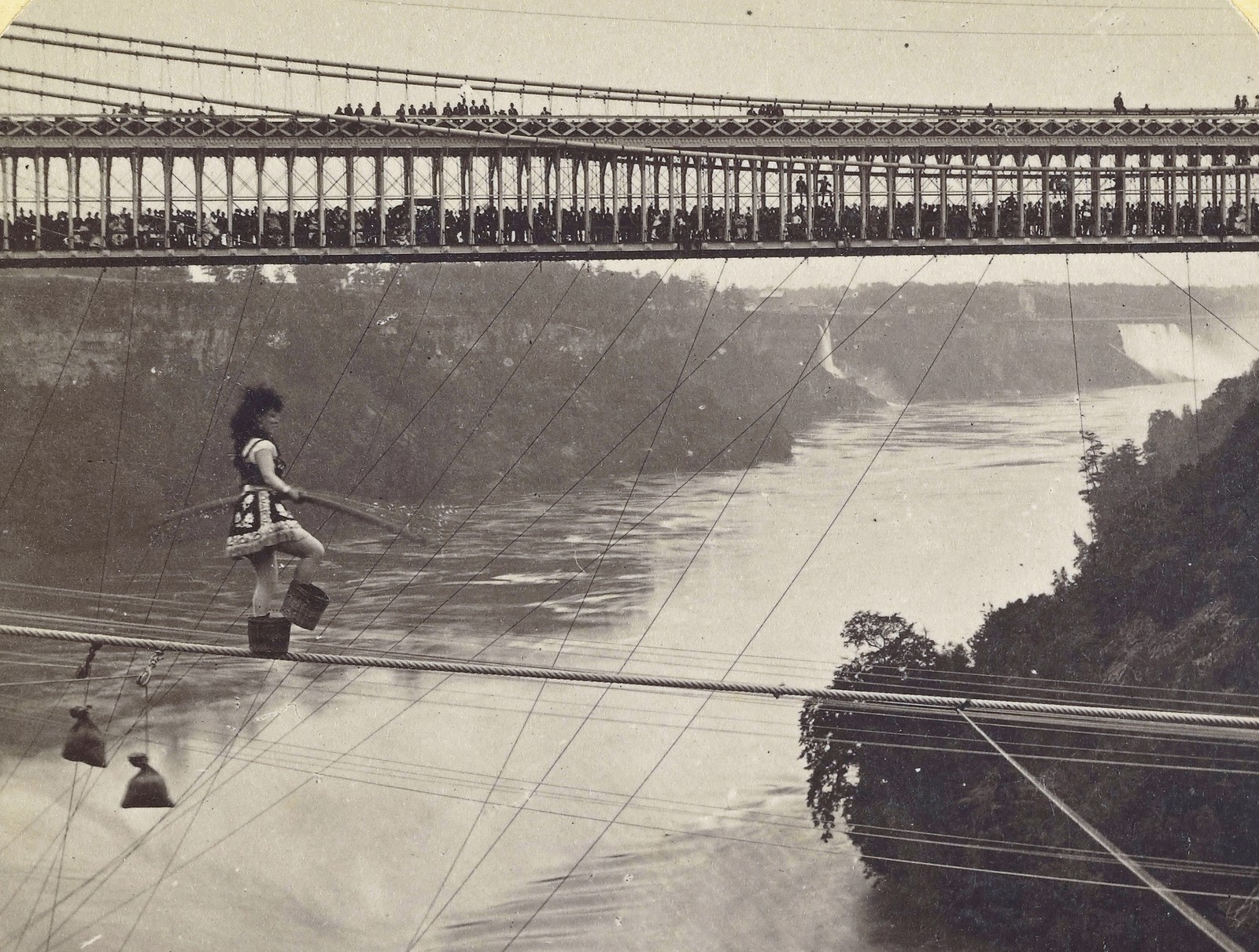 A large crowd of people look on from a bridge as a tightrope walker (wearing small baskets on their feet) walks on a rope above Niagara Falls.