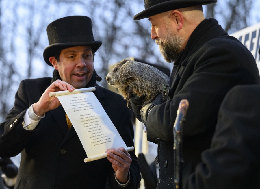 A man in a top hat holds up a proclamation to a groundhog that is being held by another man.