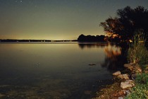 photo at night of lake shore with grassy bank, glowing horizon, and sky with bright stars