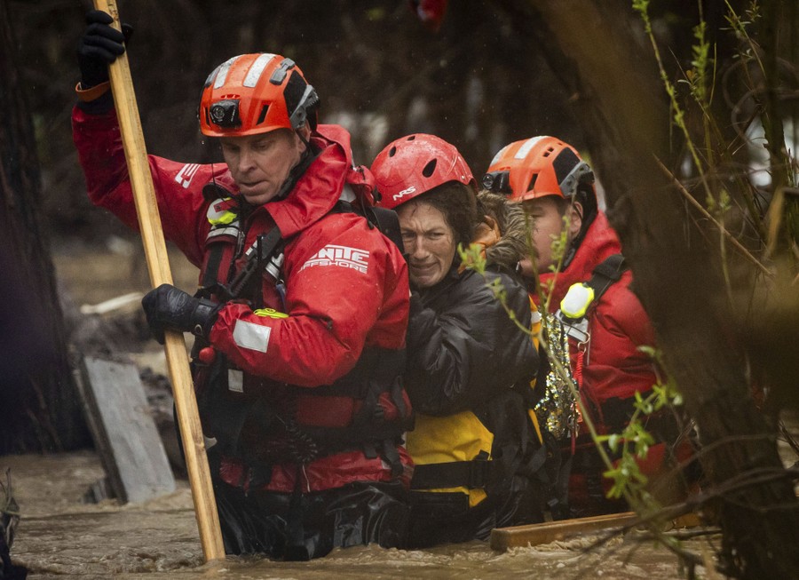 Two rescue workers help a person escape a flood, walking through waist-deep water.