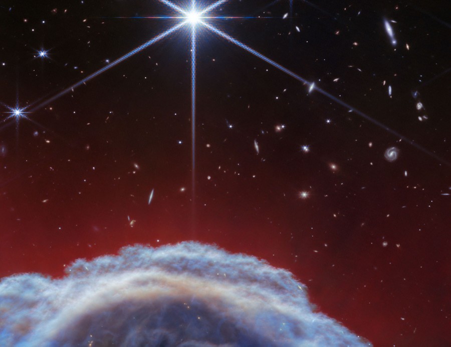 A telescope view of a cloud-like structure of gas and dust at the bottom of the image, with distant stars and galaxies visible above.