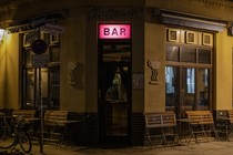 A bar at night, with an illuminated pink sign in the center and folded up tables along its windows