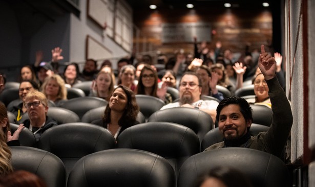 People are seated in an auditorium with rows of black leather seats. A few people raise their hands.