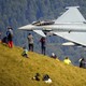 People stand on a hillside as a jet fighter flies past very closely.