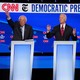 Vermont Senator Bernie Sanders, standing next to former Vice President Joe Biden and Senator Elizabeth Warren of Massachusetts, all behind lecterns on a stage, raises his hand during the fourth Democratic primary debate.