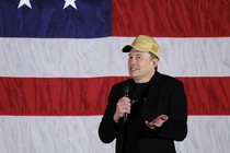 Elon Musk shrugs wearing a MAGA hat in front of an American flag