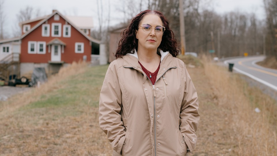 A woman wearing a brown jacket stands on the side of the road in front of a red house