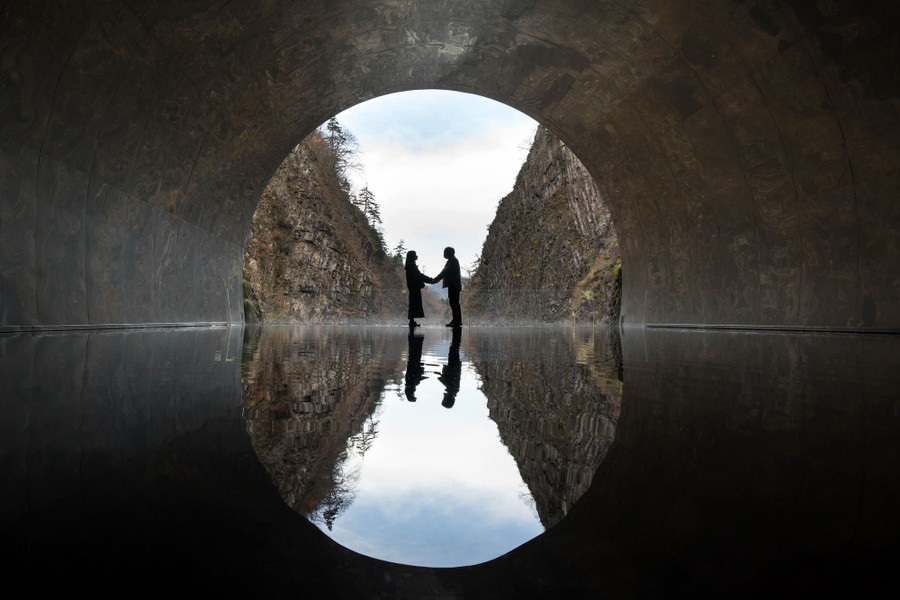 A couple poses in front of an illuminated scene of a gorge, seen inside a tunnel.