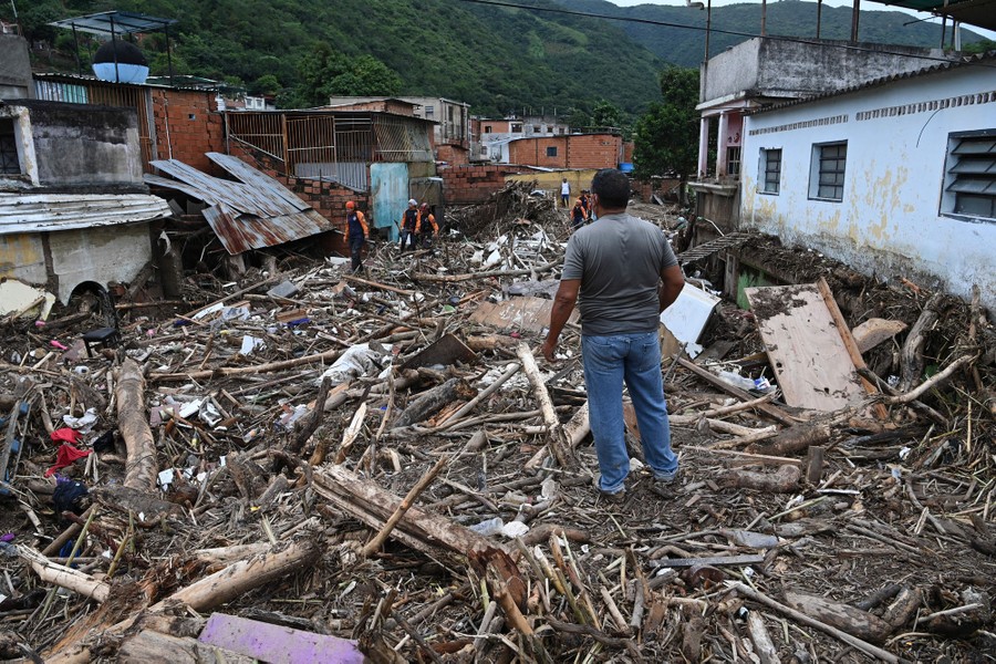 A person stands on thick piles of debris that fill a street, among damaged buildings.