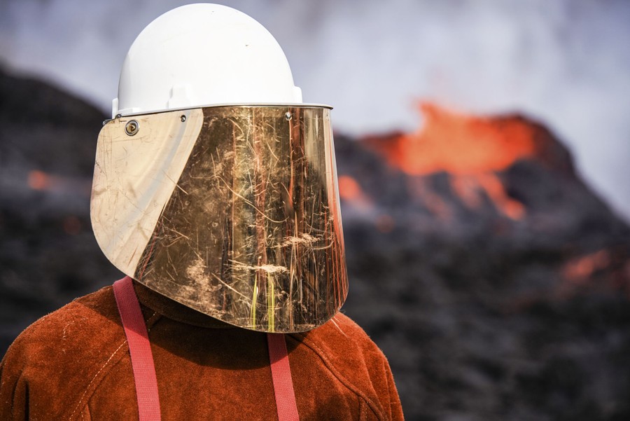 A person stands near flowing lava, wearing protective clothing and a helmet with a reflective visor.