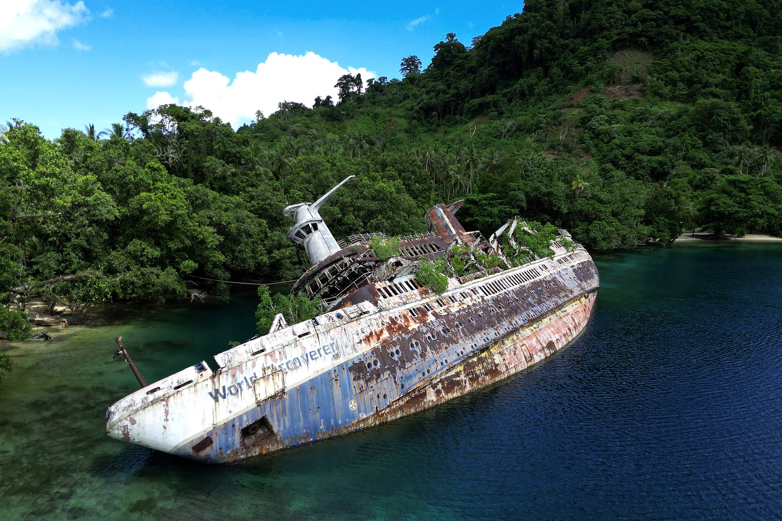 An aerial view of a rusty, wrecked, and listing cruise ship, grounded near the shore of a lush Island