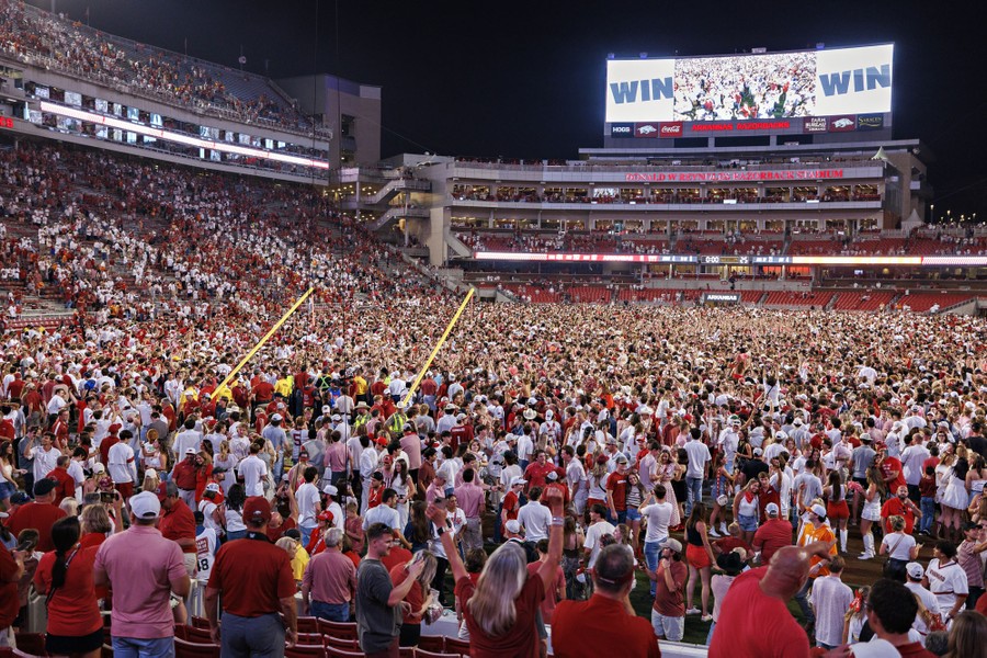The field of a football stadium is filled with fans who stormed it after a game; the goalposts seem to have been knocked over.