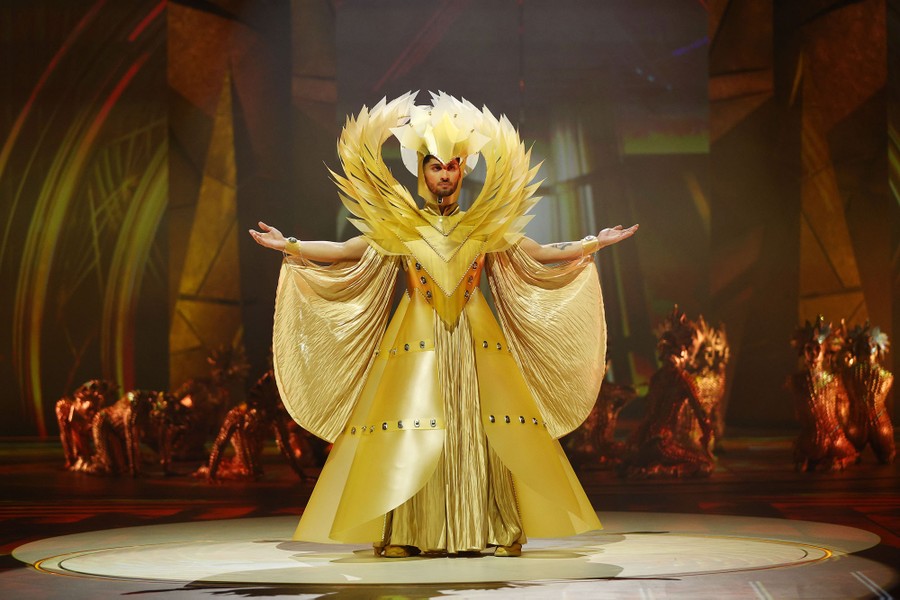 A person displays a golden costume with a feathery crest stretching from their shoulders to above their head.