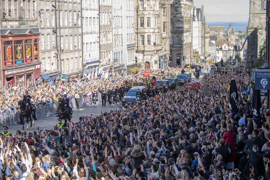 A large crowd stands on either side of a street as a procession of cars follows a hearse carrying Queen Elizabeth's coffin.