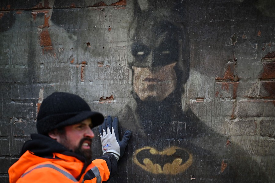 A person leans against a prop wall with a mural of Batman painted on it.