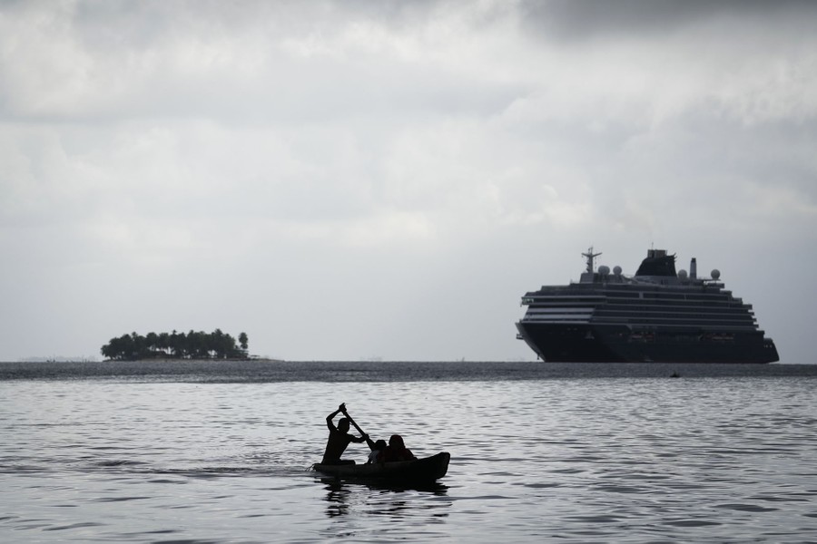 Several people in a small boat paddle by, with a huge cruise ship visible in the distance.