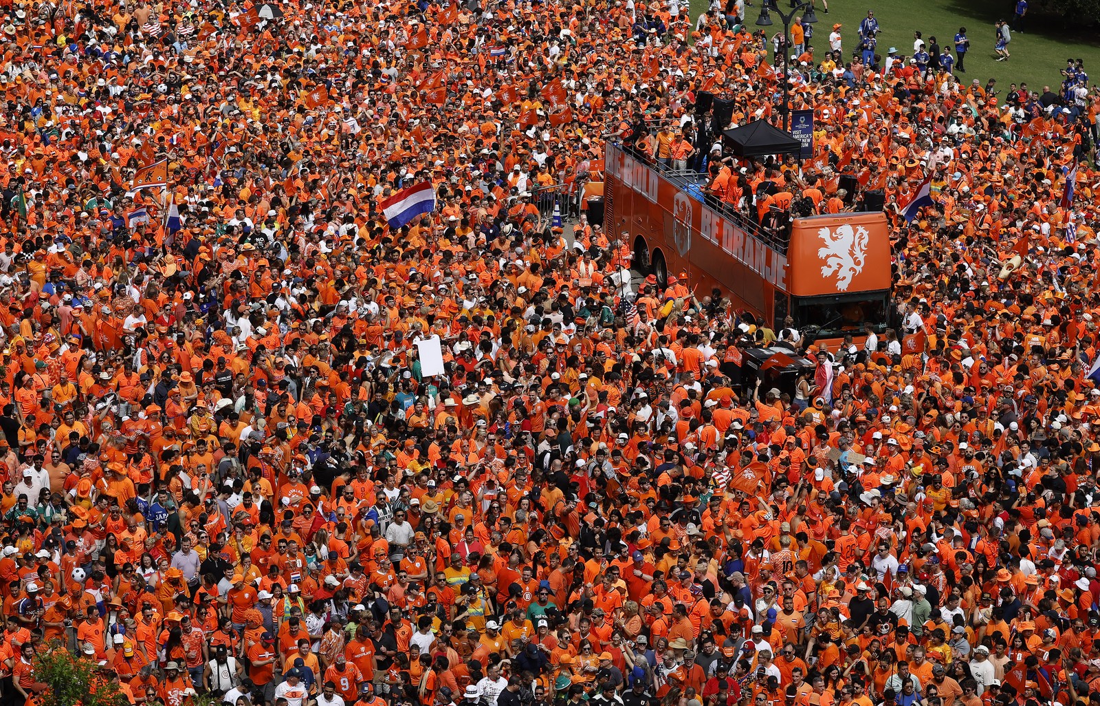 An elevated view of a large crowd of soccer fans, almost all wearing orange, gathered around a double-decker bus