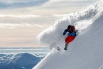Photograph of a skier in red pants, a blue jacket, and a black helmet carving a turn in deep powder on a steep slope, with mountains in the background