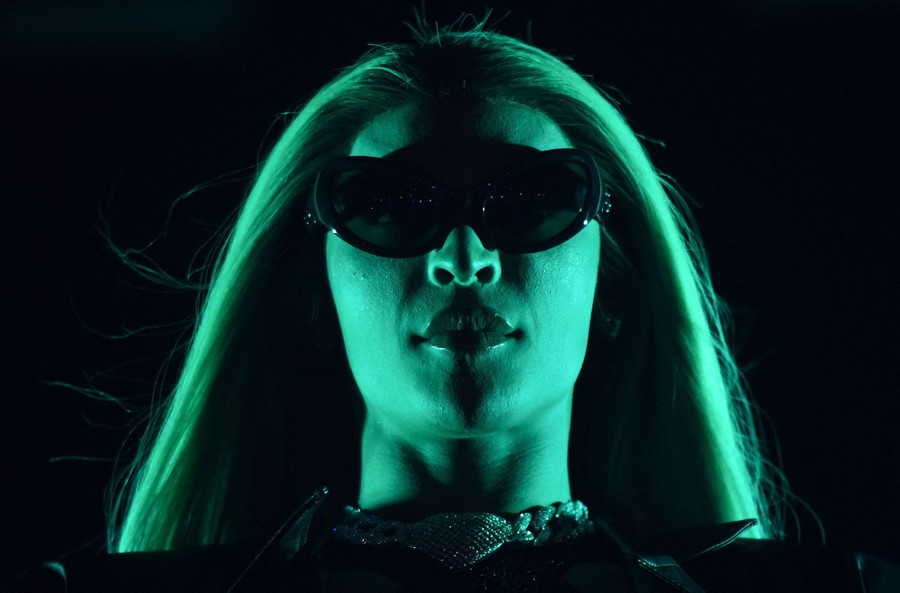 A close view of a performer with long hair and dark glasses, lit by green light.