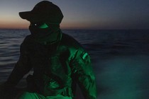 Photograph of a masked soldier on a small boat at dusk, with a green light shining on him