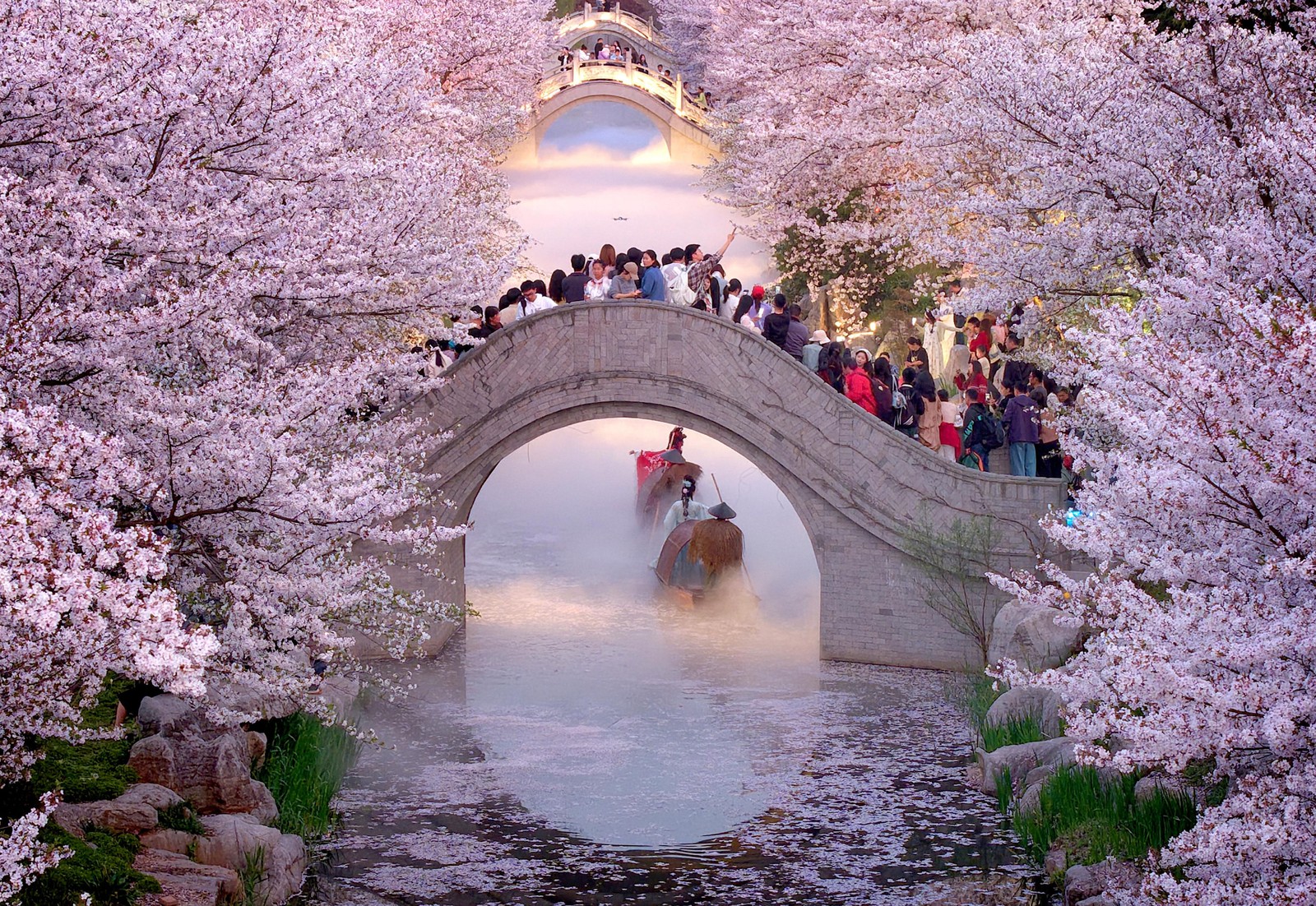Visitors stand on an arched bridge above a canal, surrounded by blooming trees, watching costumed people row boats along a fog-covered canal below.