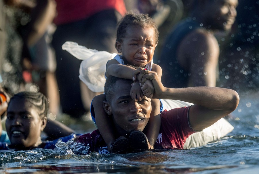 A person carries a small upset child on their shoulders as they cross a chest-deep river.