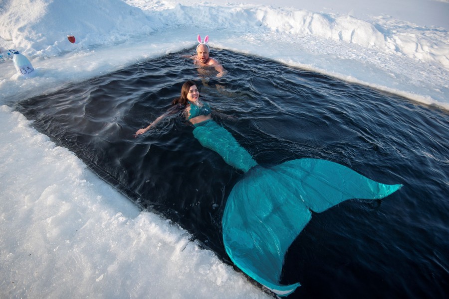 A man wearing bunny ears and a woman dressed as a mermaid swim in a rectangular hole cut into lake ice.