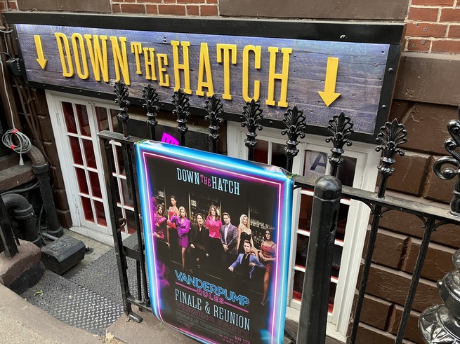 A sign advertising a Vanderpump Rules finale watch party, in front of a garden-level dive bar called Down the Hatch.