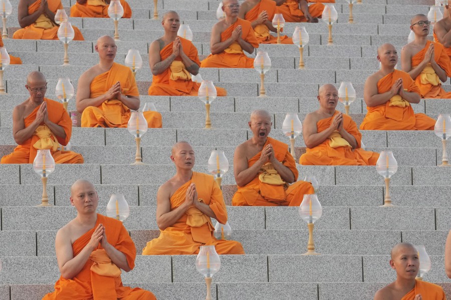 Buddhist monks in orange robes sit in rows, cross-legged on stone steps, praying.