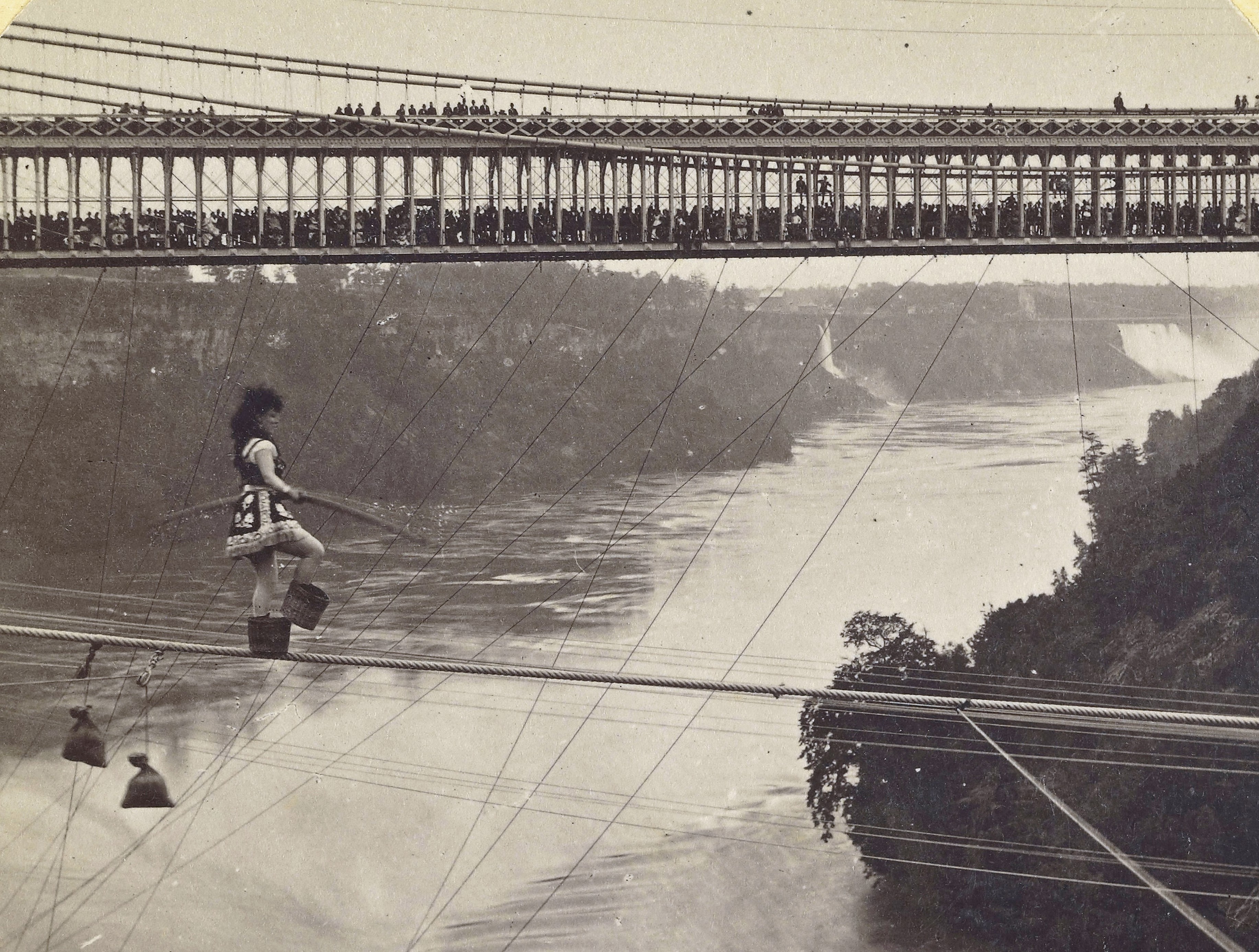 A large crowd of people look on from a bridge as a tightrope walker (wearing small baskets on their feet) walks on a rope above Niagara Falls.