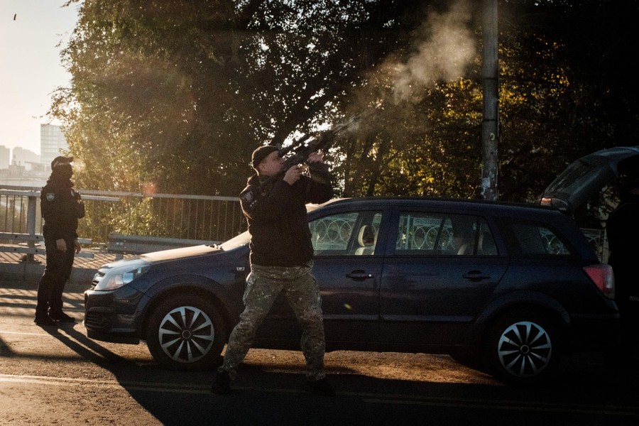 A police officer standing in a street fires his weapon at an upward angle.