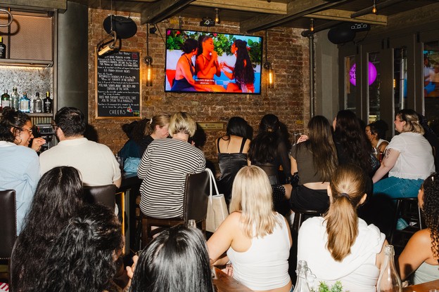 A group of adults watching "Love Island USA" in a bar with their backs turned toward the camera