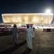 A photo of people walking through metal gates toward a lit-up soccer stadium shaped like an elongated basket