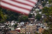 Color photograph of the town of Charleroi seen at a distance on what appears to be an overcast day. A blurry American flag whips across the top of the frame at close range.