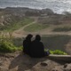 A color photograph of two women dressed in black sitting on a cliff face overlooking the water in Beirut.