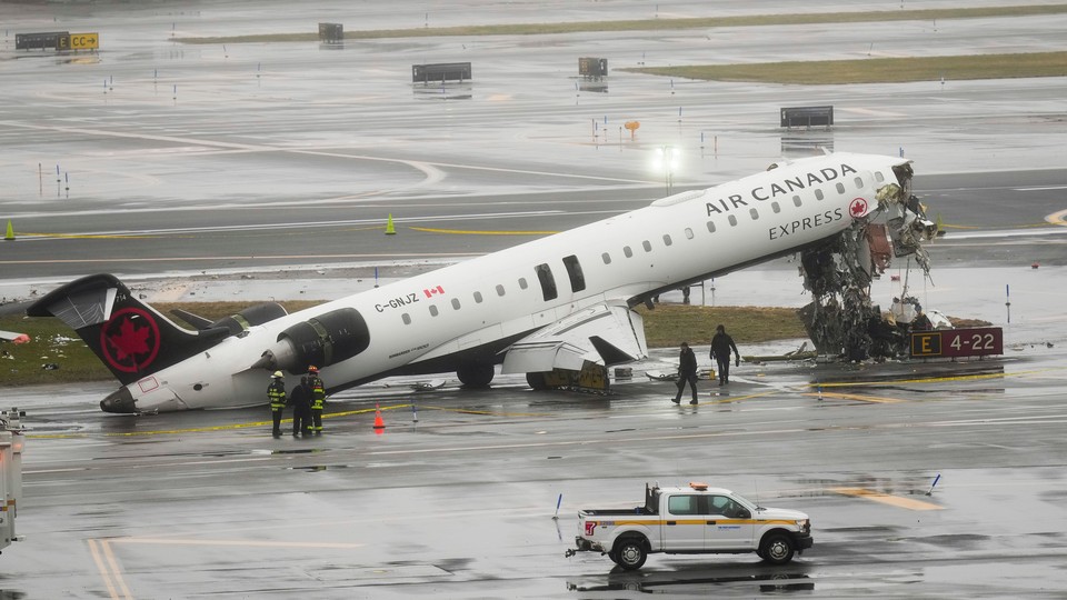 Photograph of the wreckage of Air Canada airplane on the runway of an airport, with the plane’s nose upturned with exposed wiring and torn metal exposing its inside