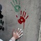 Photograph of a one green and one red handprint on a concrete wall above a bystander with a white-painted hand leaving a third imprint. The photo is surrounded by a thick black border.