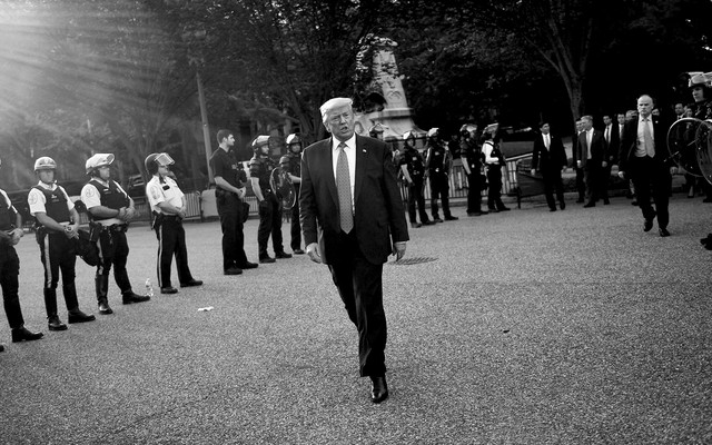 Donald Trump, with police officers behind him, walks outside the White House. The photo is black and white.