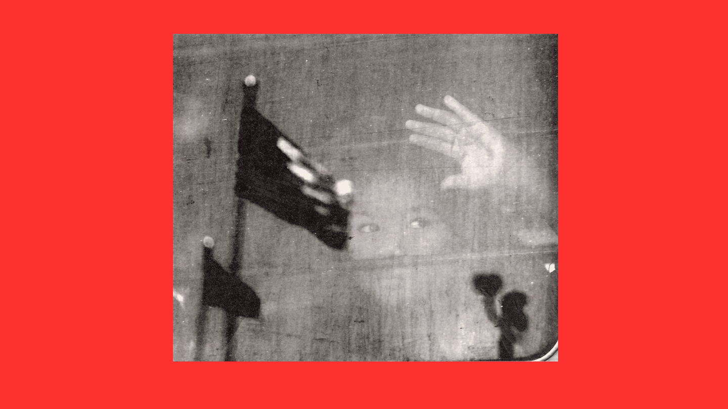 fotografía en blanco y negro de una joven que mira por una ventana y presiona su mano contra ella con el reflejo de dos banderas estadounidenses