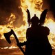 A person wearing a fanciful Viking costume, holding an axe, stands in front of a burning replica of a viking ship, seen in silhouette.
