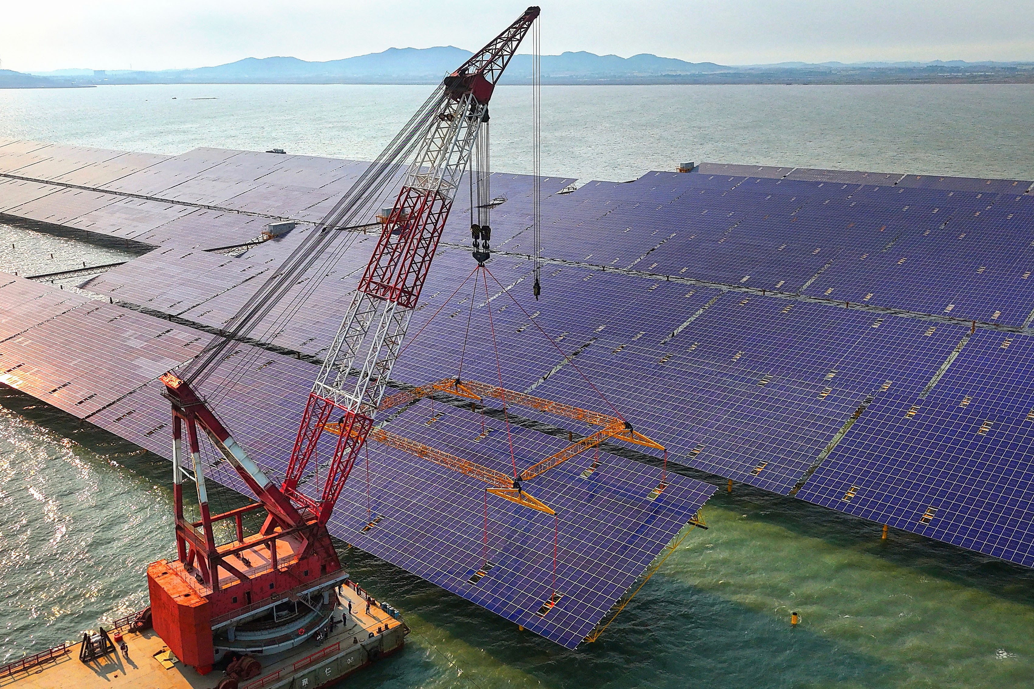 A crane on a floating barge positions a large solar panel for installation.