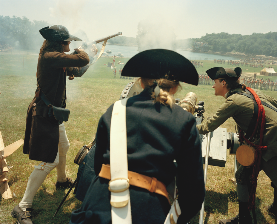 photo of reenactment of battle with American soldiers in period clothing, one firing a musket and two aiming a cannon toward the British lines, with the shore and spectators in the background